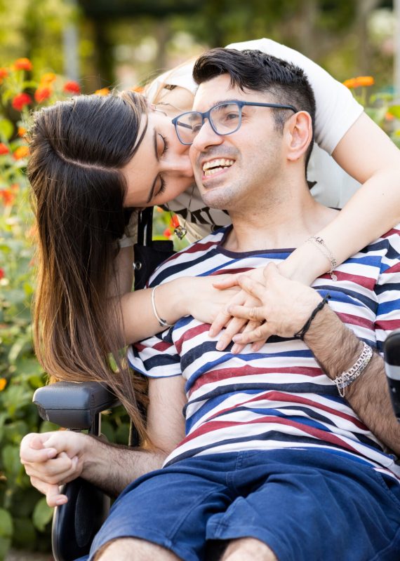 An adult man with a disability sits in an automatic wheelchair with a young woman in a park at sunset.The woman kisses the disabled man on the face.Unconditional support for people with disabilities.